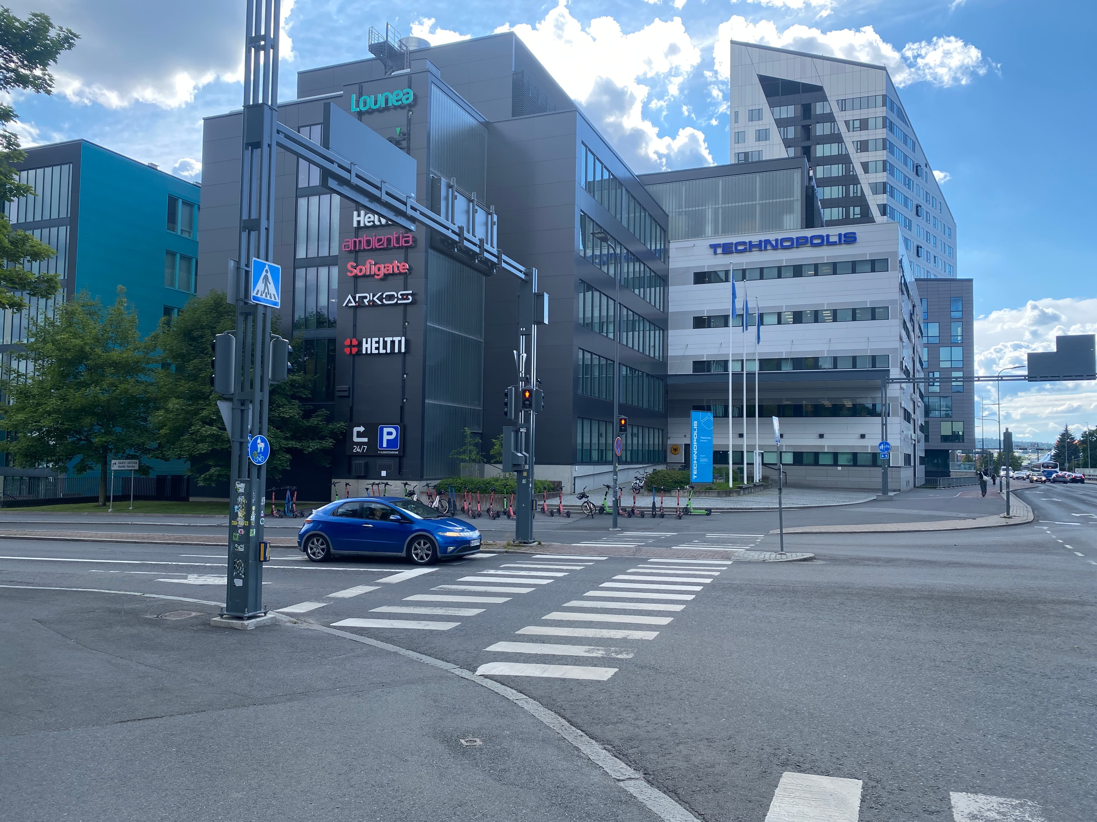 Näkymä kaupunkiristeyksestä, jossa on moderneja toimistorakennuksia ja suojatietä ylittävä sininen auto.