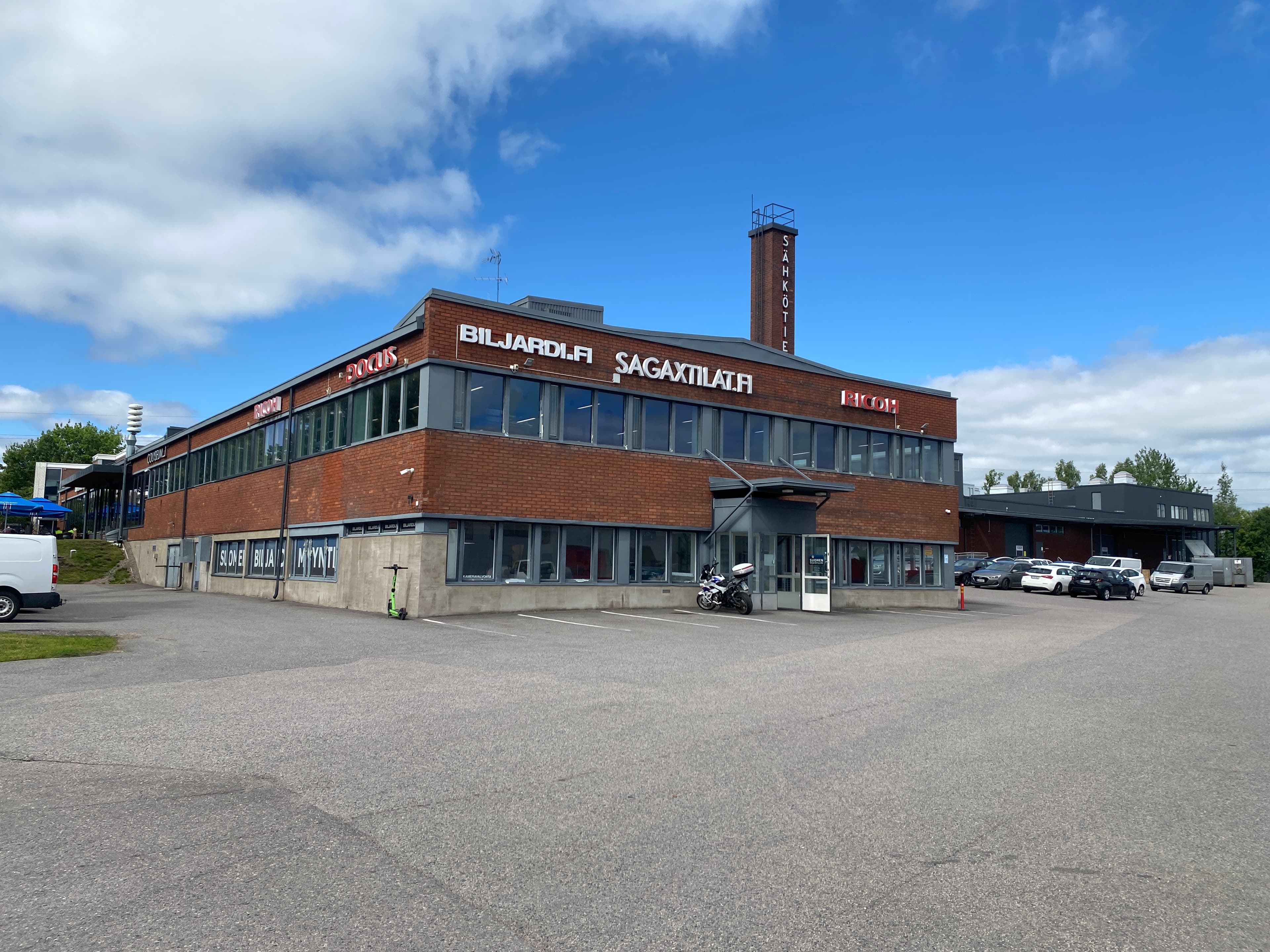 Kaksikerroksinen punatiilinen liikerakennus, jonka julkisivussa on useita yrityskylttejä, asfaltoidun pysäköintialueen reunalla aurinkoisena päivänä.