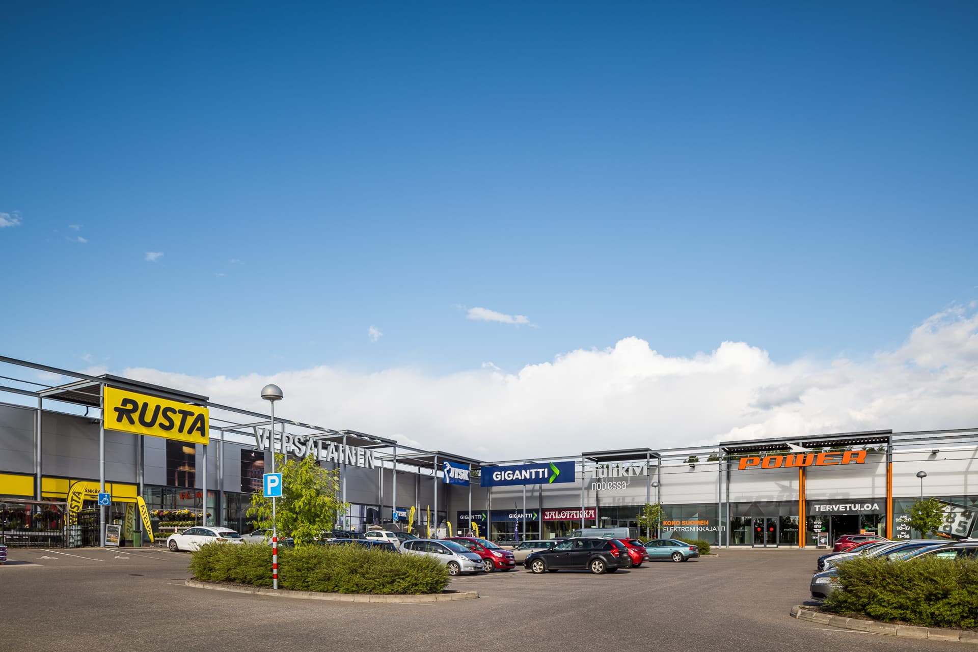 Kauppakeskuksen pysäköintialue ja liikerakennuksia aurinkoisena päivänä.