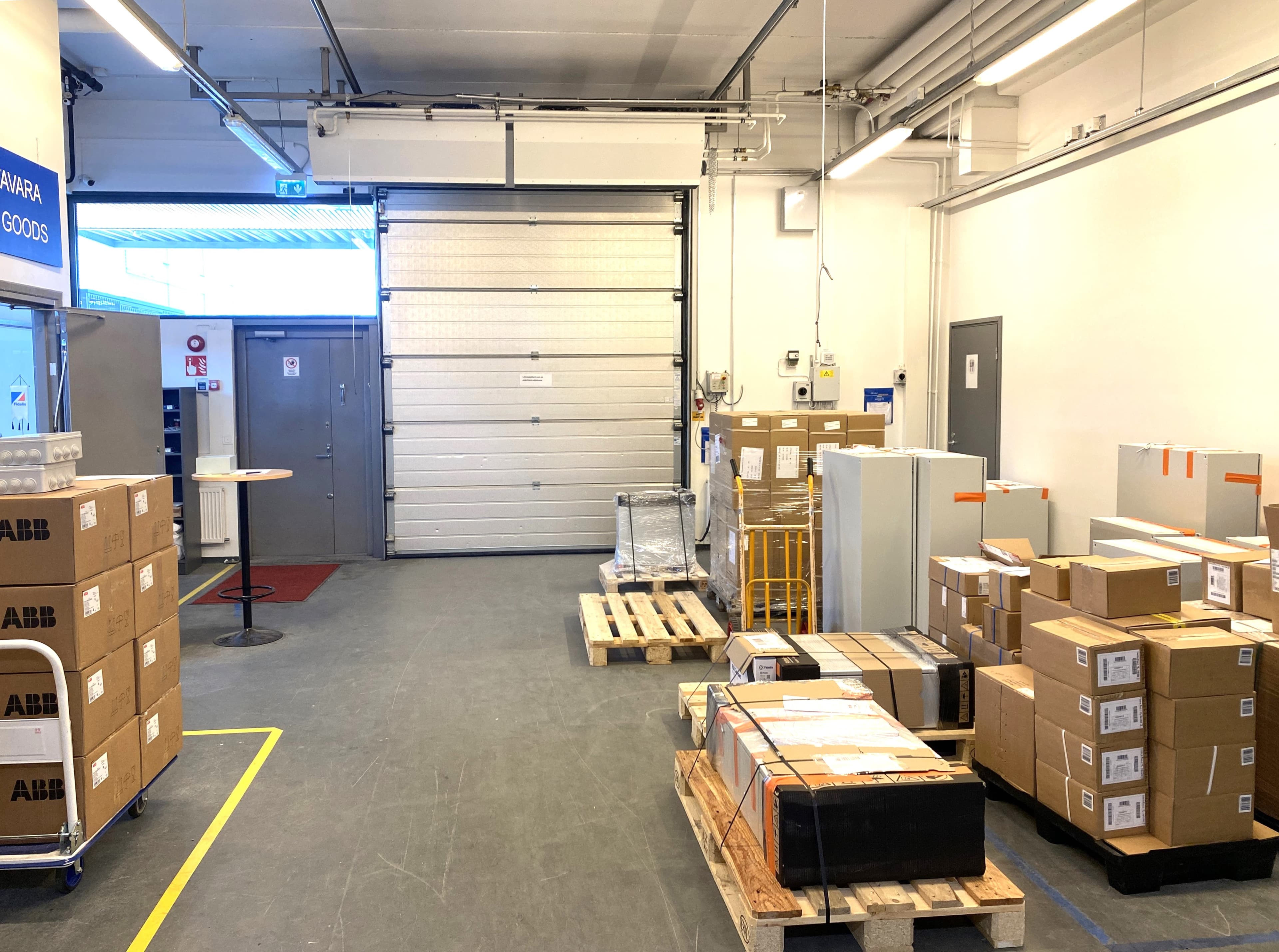 Varaston lastausalue, jossa on useita tavaralaatikoita, kuormalavoja ja suuri teollisuusovi.