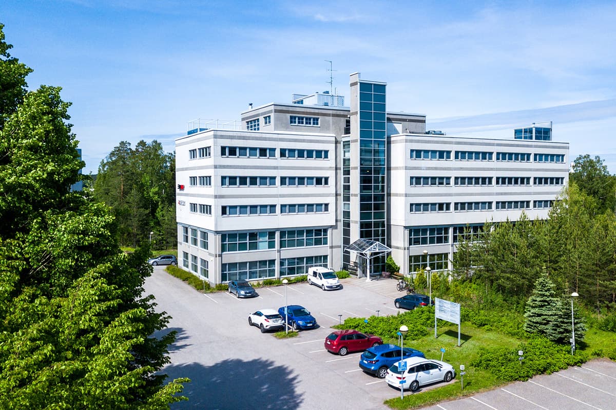 Monikerroksinen vaalea toimistorakennus ja sen edessä oleva parkkipaikka aurinkoisena päivänä.