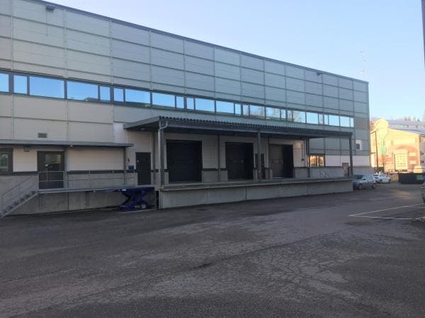 Suuri vaaleanharmaa teollisuusrakennus, jonka edustalla on katettu lastauslaituri nosto-ovineen ja asfaltoitu piha.