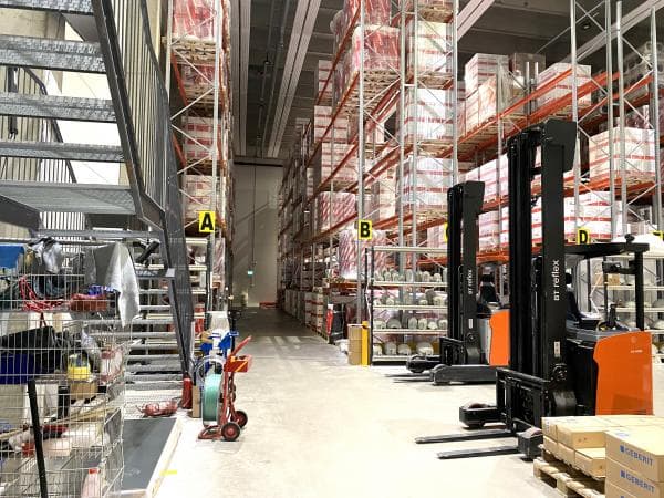 Varastohalli, jossa on korkeita hyllyjä, trukkeja ja metalliportaat.
