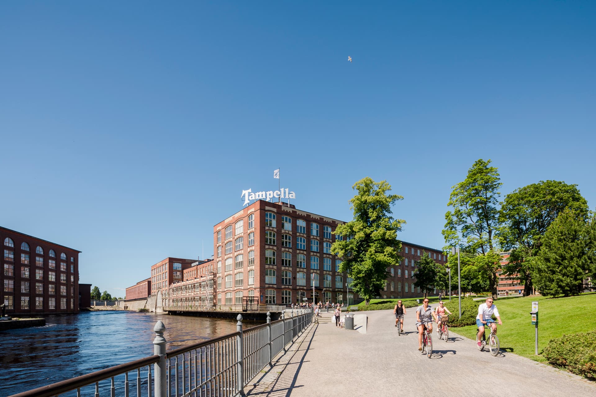 Ihmisiä pyöräilemässä ja kävelemässä Tammerkosken rannalla sijaitsevalla tiellä, taustalla Tampellan punatiilisiä tehdasrakennuksia.