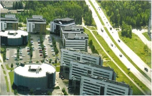 Ilmakuva modernista toimistokeskittymästä, jossa on useita rakennuksia, laaja pysäköintialue ja maantie metsäisessä ympäristössä.