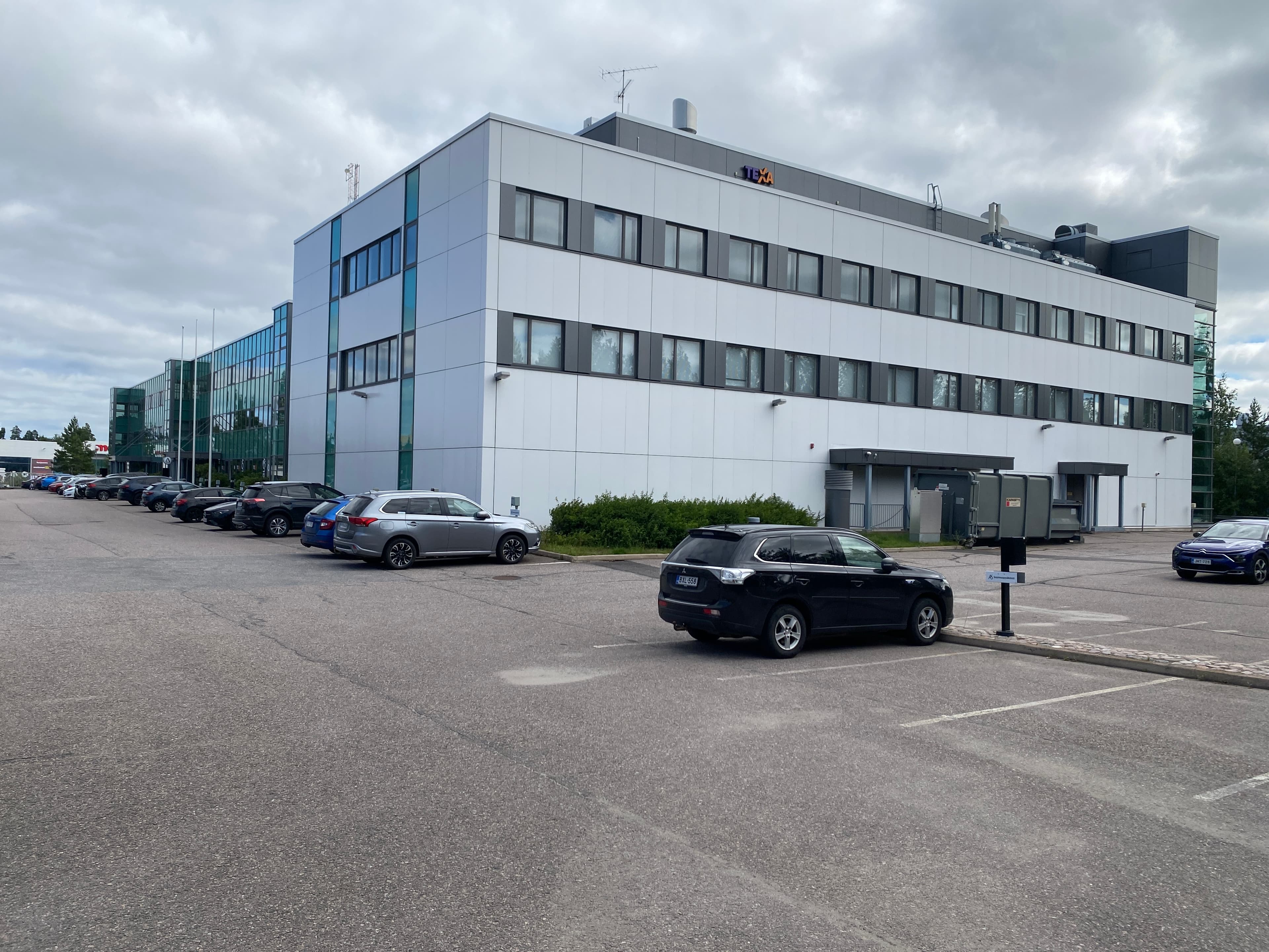 Valkoinen ja harmaa moderni toimistorakennus, jonka edessä on pysäköityjä autoja asfaltoidulla parkkipaikalla.