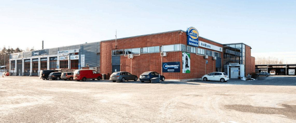 Liikerakennus, jossa sijaitsee useita autohuoltoyrityksiä, ja autoja pysäköitynä sen edustalla.