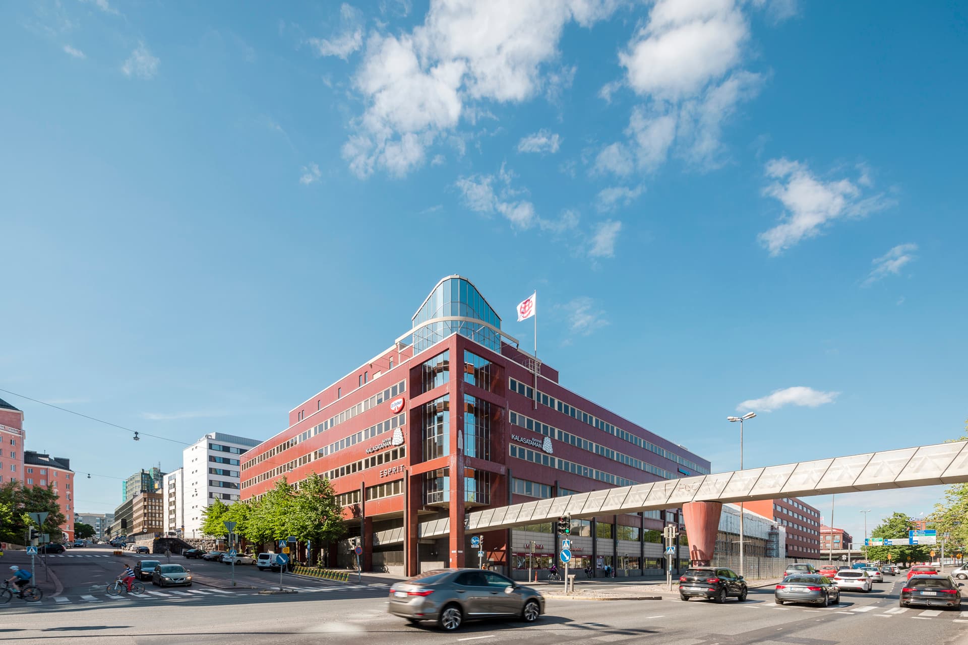 Aurinkoinen kaupunkinäkymä, jossa on suuri punatiilinen rakennus, vilkas risteys ja jalankulkusilta.