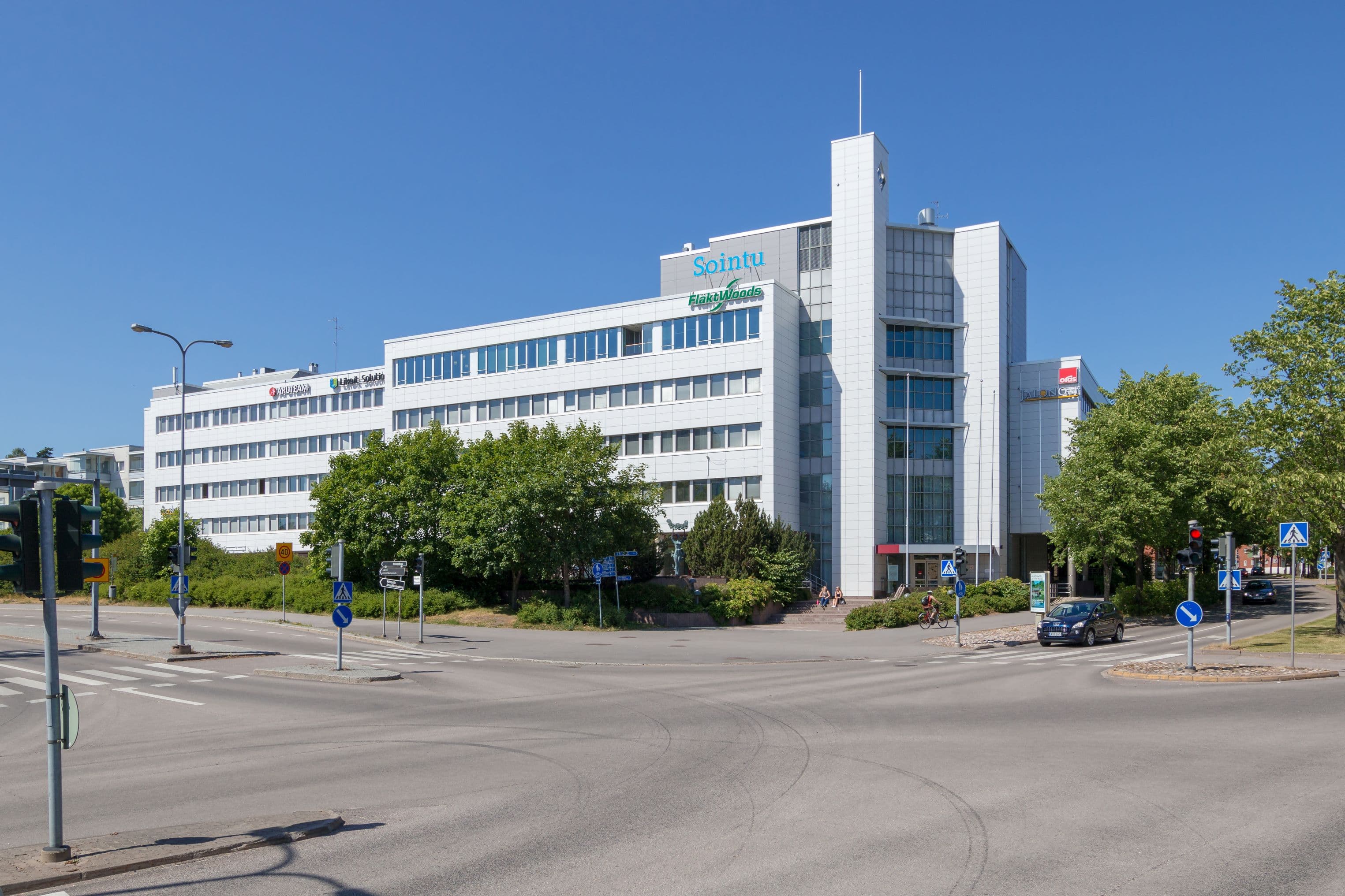 Valkoinen toimistorakennus katujen risteyksessä aurinkoisena päivänä.