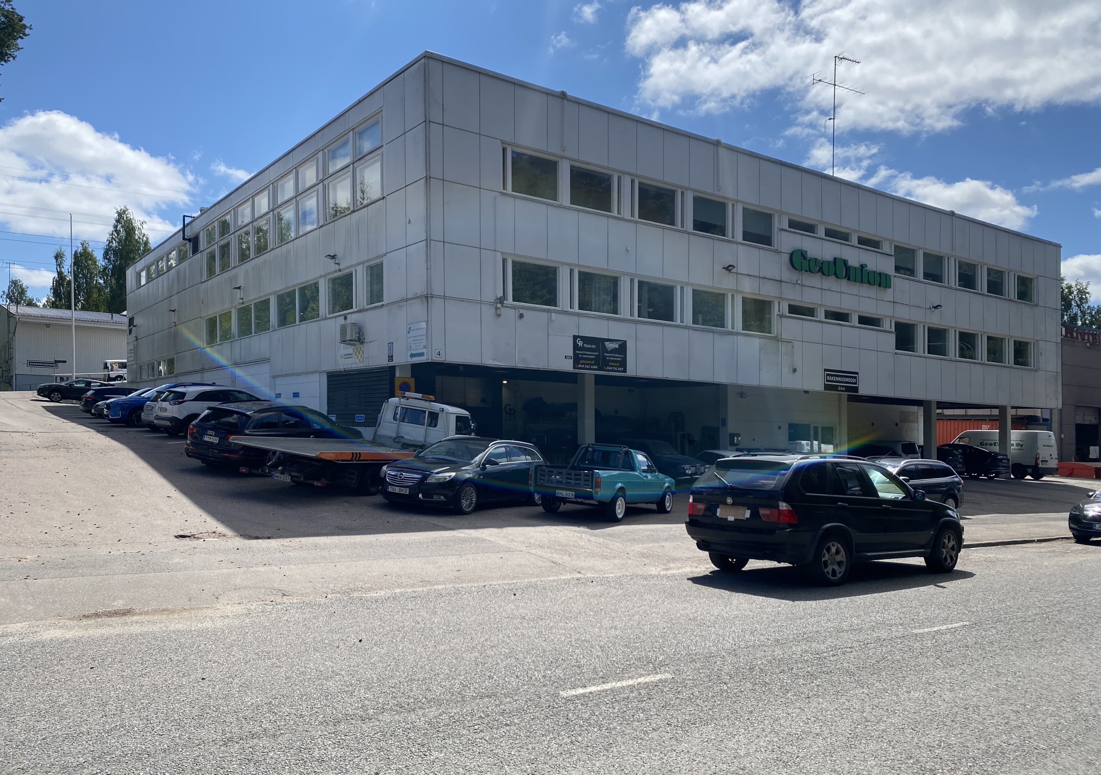 Valkoinen liikerakennus ja sen edustalle pysäköityjä autoja aurinkoisena päivänä.