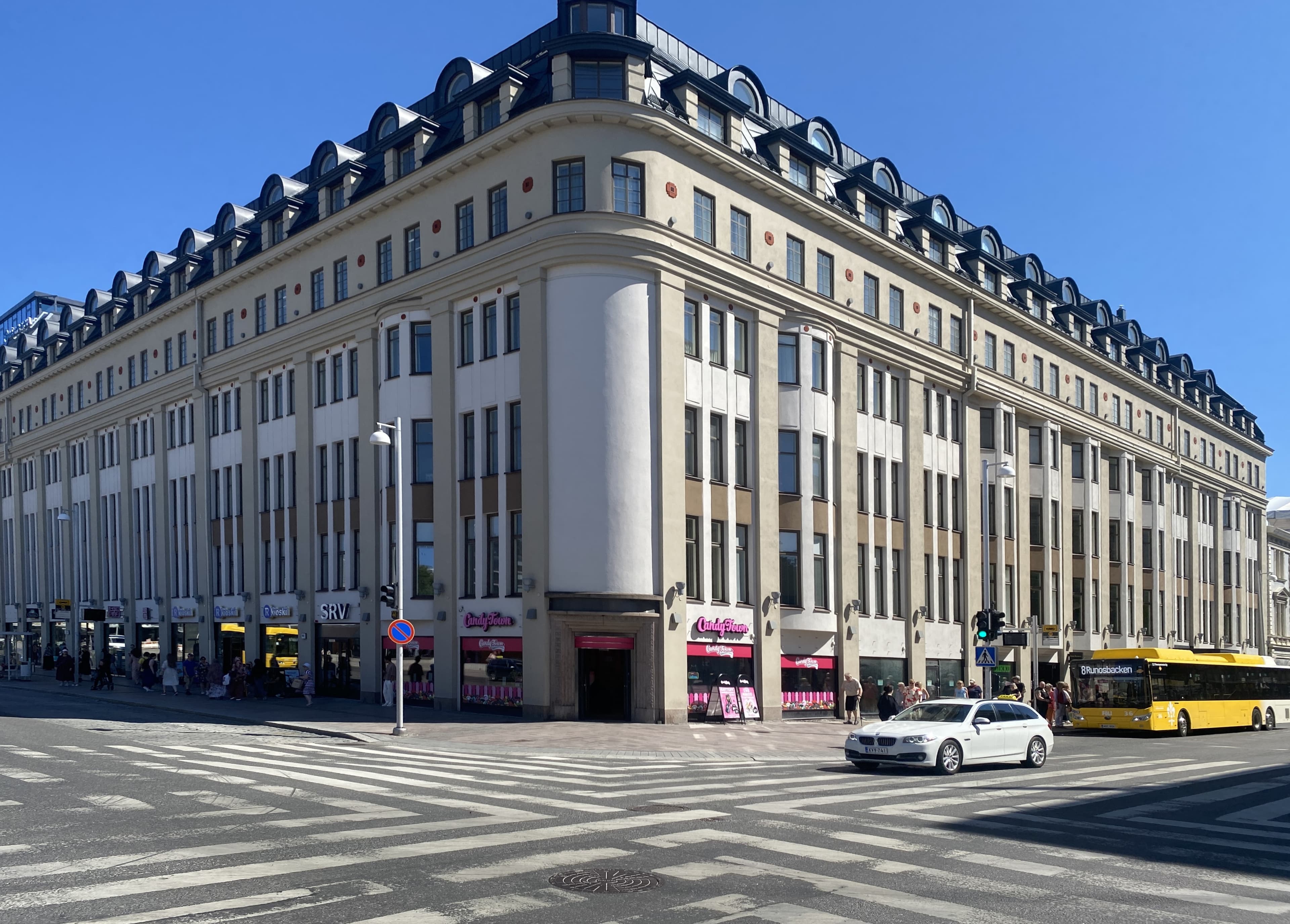 Suuri vaalea kulmarakennus, valkoinen auto ja keltainen bussi kaupungin keskustassa aurinkoisena päivänä.