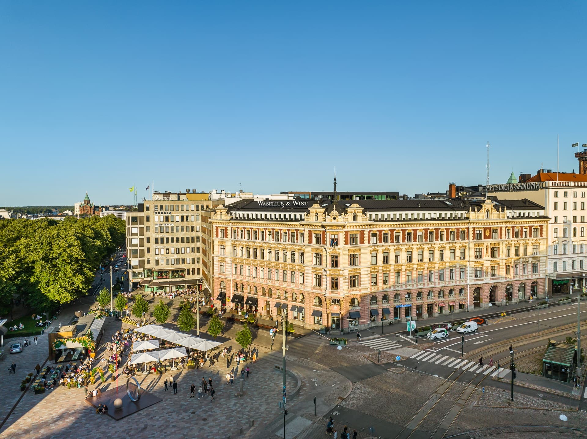 Ilmakuva Helsingin keskustasta, jossa näkyy historiallisia rakennuksia, Esplanadin puisto ja ihmisiä aukiolla aurinkoisena päivänä.