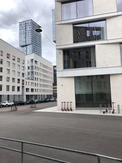 Moderni kaupunkinäkymä, jossa on vaaleita rakennuksia, sähköpotkulautoja ja korkea tornitalo taustalla.