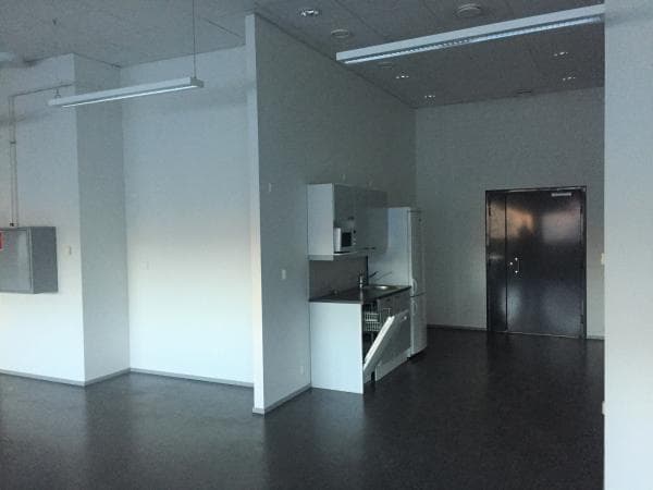Pelkistetty ja avoin sisätila, jossa on pieni keittiönurkkaus, valkoiset seinät ja tummanharmaa lattia.