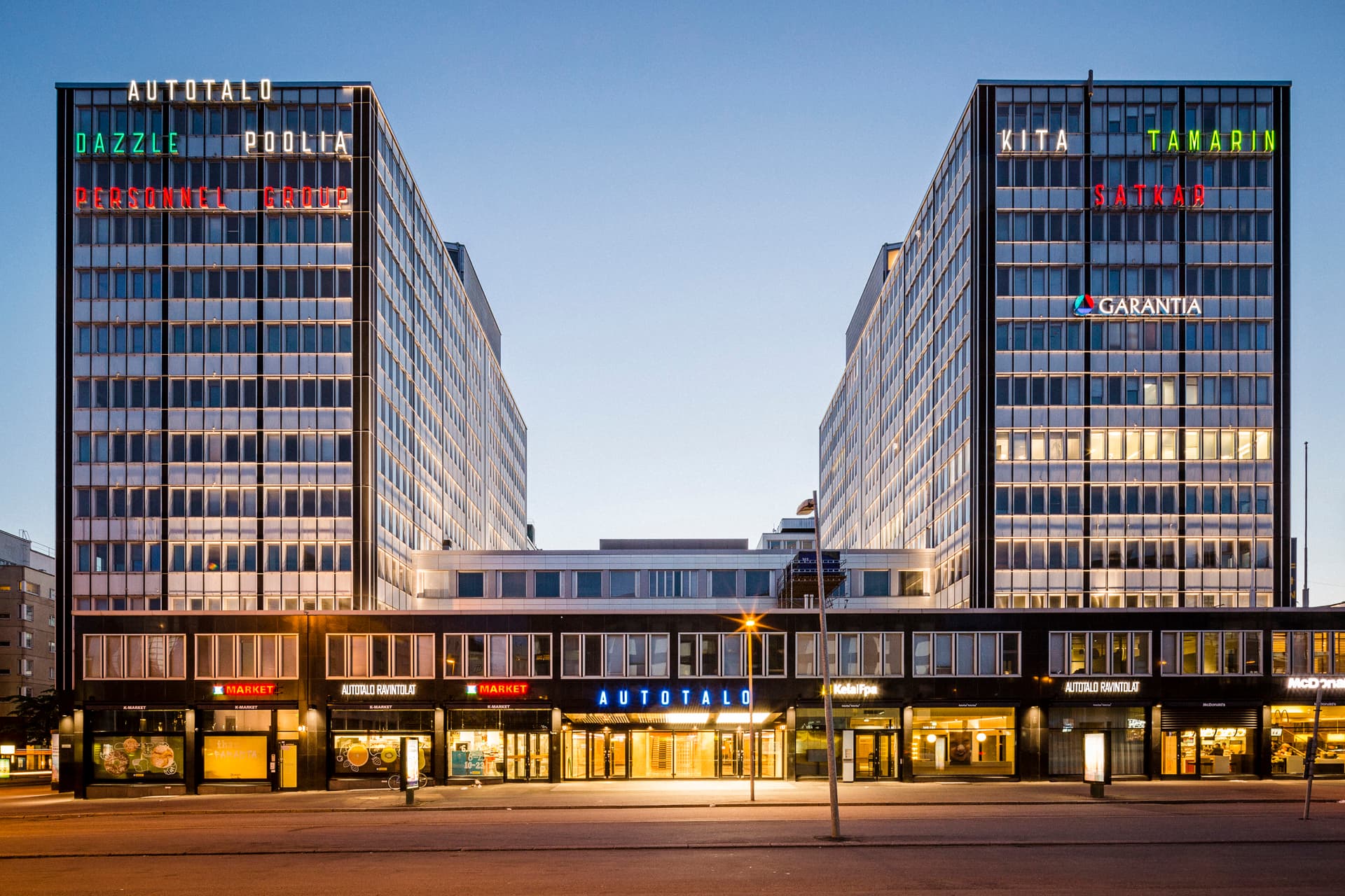 Autotalo-rakennus ja katutason liiketiloja Helsingissä iltavalaistuksessa.