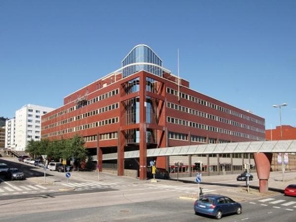 Punainen toimistorakennus ja sen ohi kulkeva kävelysilta aurinkoisessa kaupunkiympäristössä.