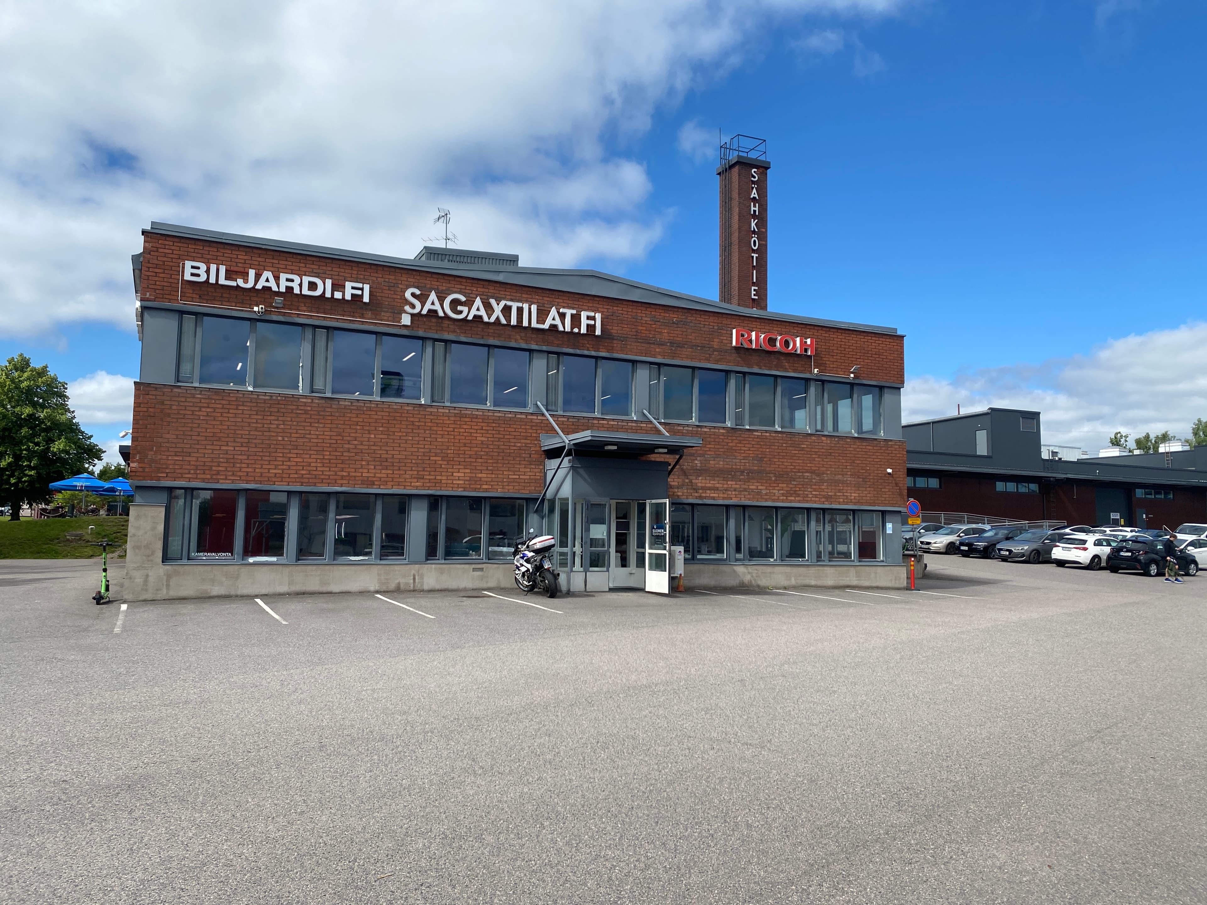 Kaksikerroksinen tiilirakennus, jonka julkisivussa on useita yrityskylttejä ja jonka edessä on asfaltoitu pysäköintialue.