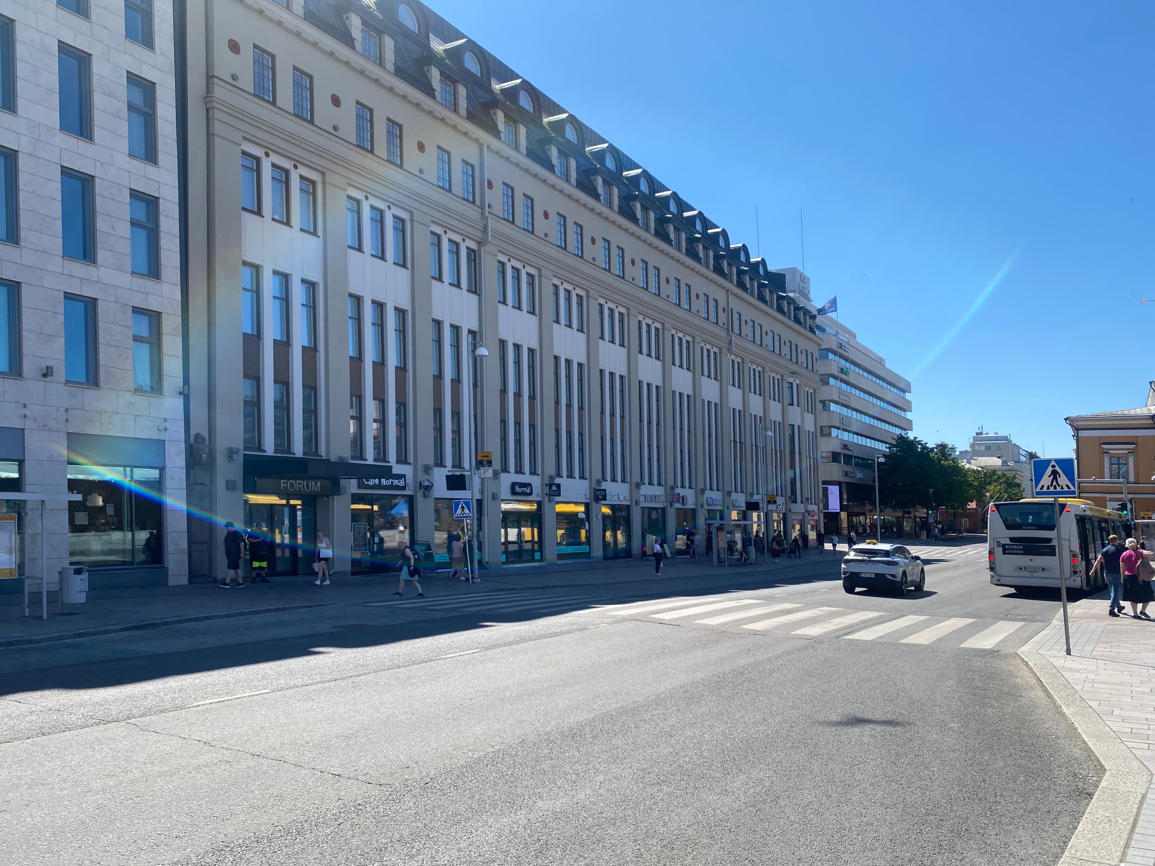 Aurinkoinen katunäkymä, jossa on suuria rakennuksia, liiketiloja, auto ja bussi.