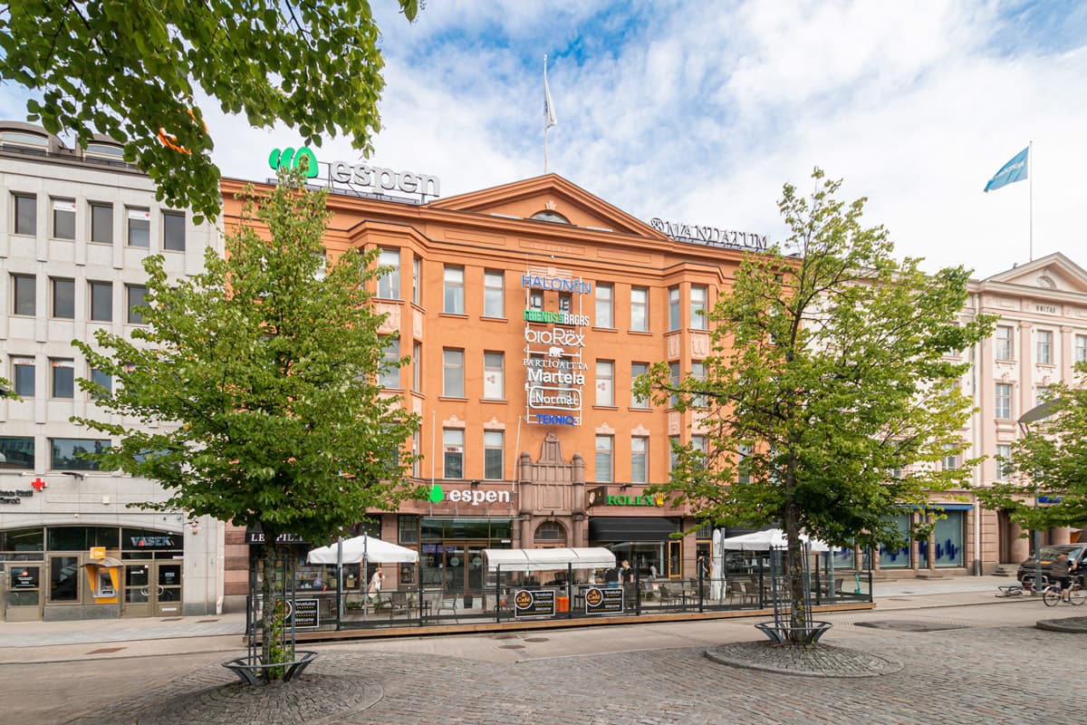 Oranssi kauppakeskusrakennus ja sen edustalla olevat puut sekä terassi aurinkoisena päivänä.
