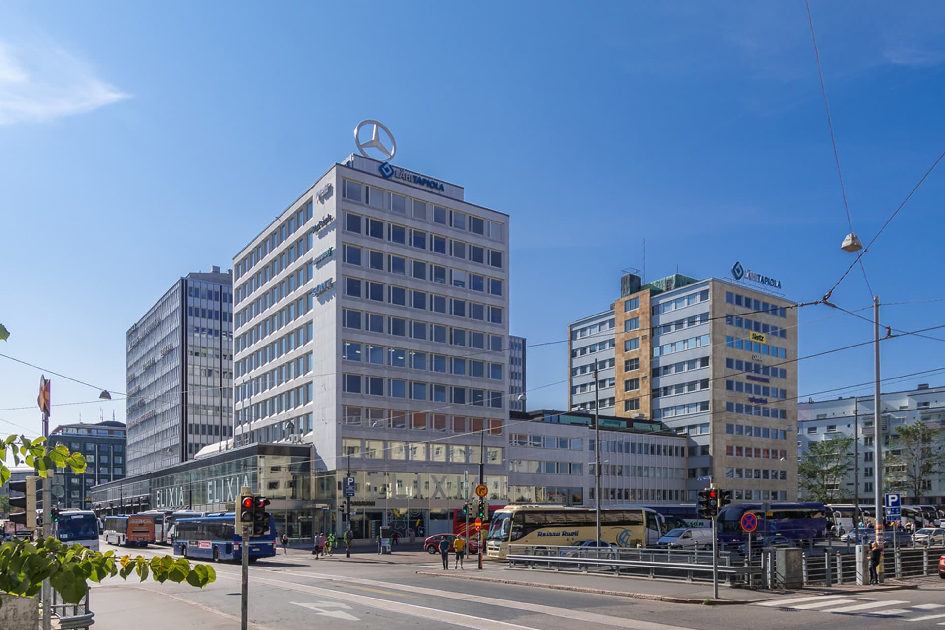 Kaupunkinäkymä, jossa on korkeita toimistorakennuksia, busseja ja liikennettä aurinkoisena päivänä.