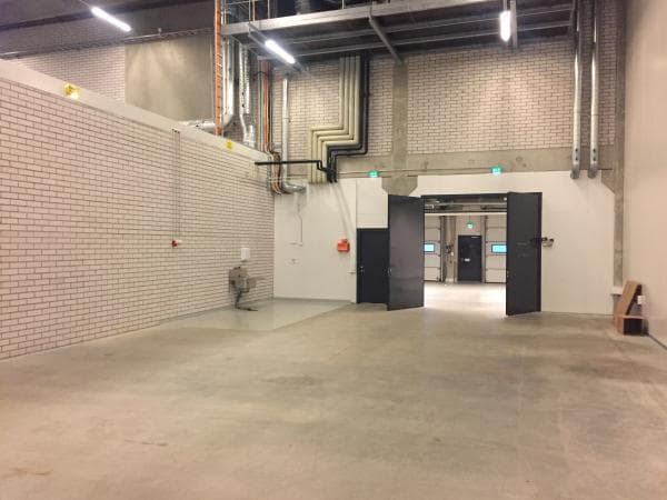 Avara ja tyhjä teollisuushalli, jossa on valkoinen tiiliseinä, harmaa betonilattia ja näkyviä kattorakenteita sekä putkistoja. Taustalla näkyvät avoimet pariovet toiseen tilaan.