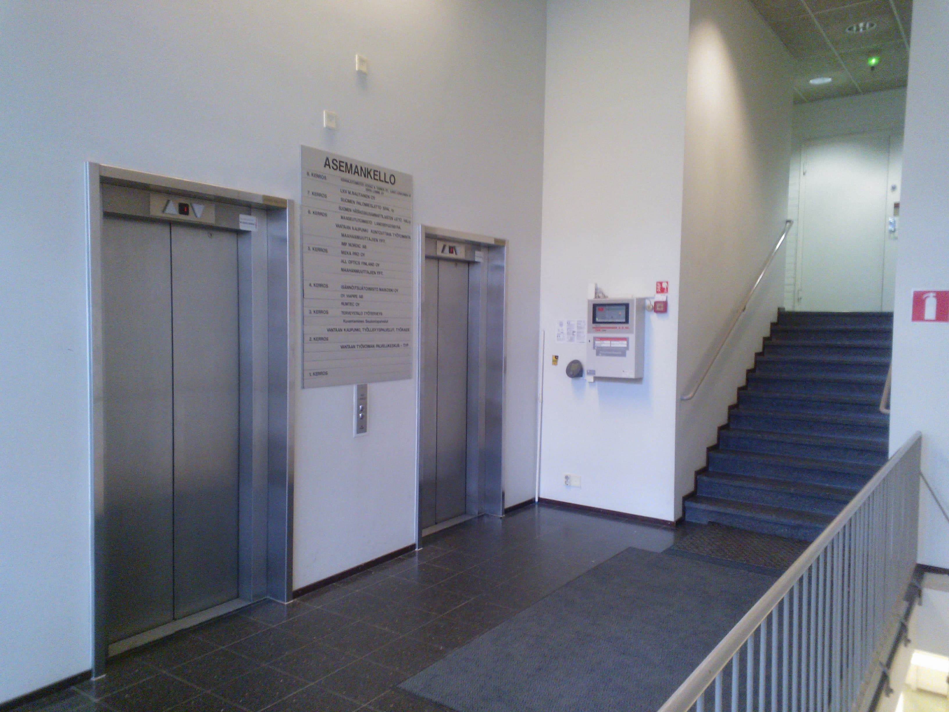 Rakennuksen aula, jossa on kaksi hissiä, opastaulu ja portaat.