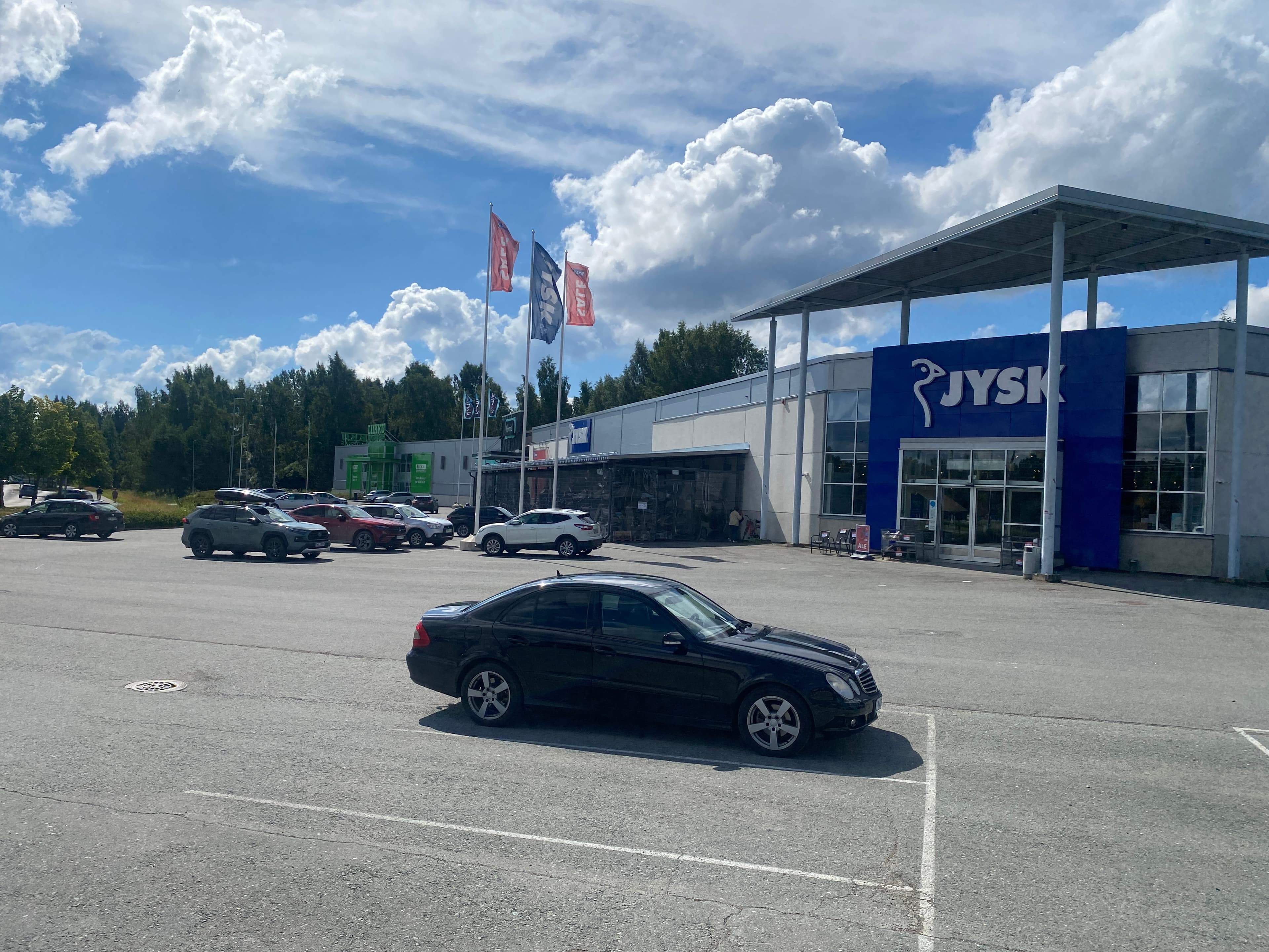 Musta auto pysäköitynä JYSK-myymälän edustalla olevalla suurella parkkipaikalla aurinkoisena päivänä.