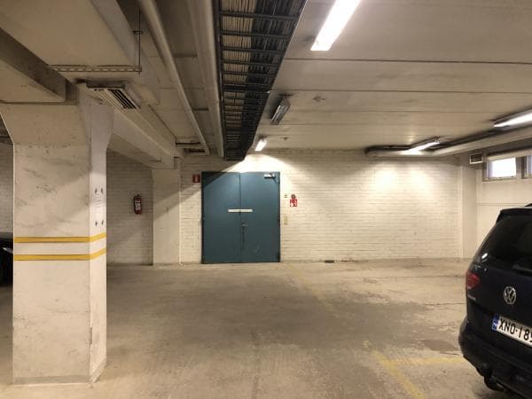 Näkymä sisäpysäköintihallista, jossa on etualalla betonipilari keltaisilla raidoilla, taustalla valkoinen tiiliseinä ja siniset pariovet sekä oikeassa reunassa osa tummansinisestä autosta.