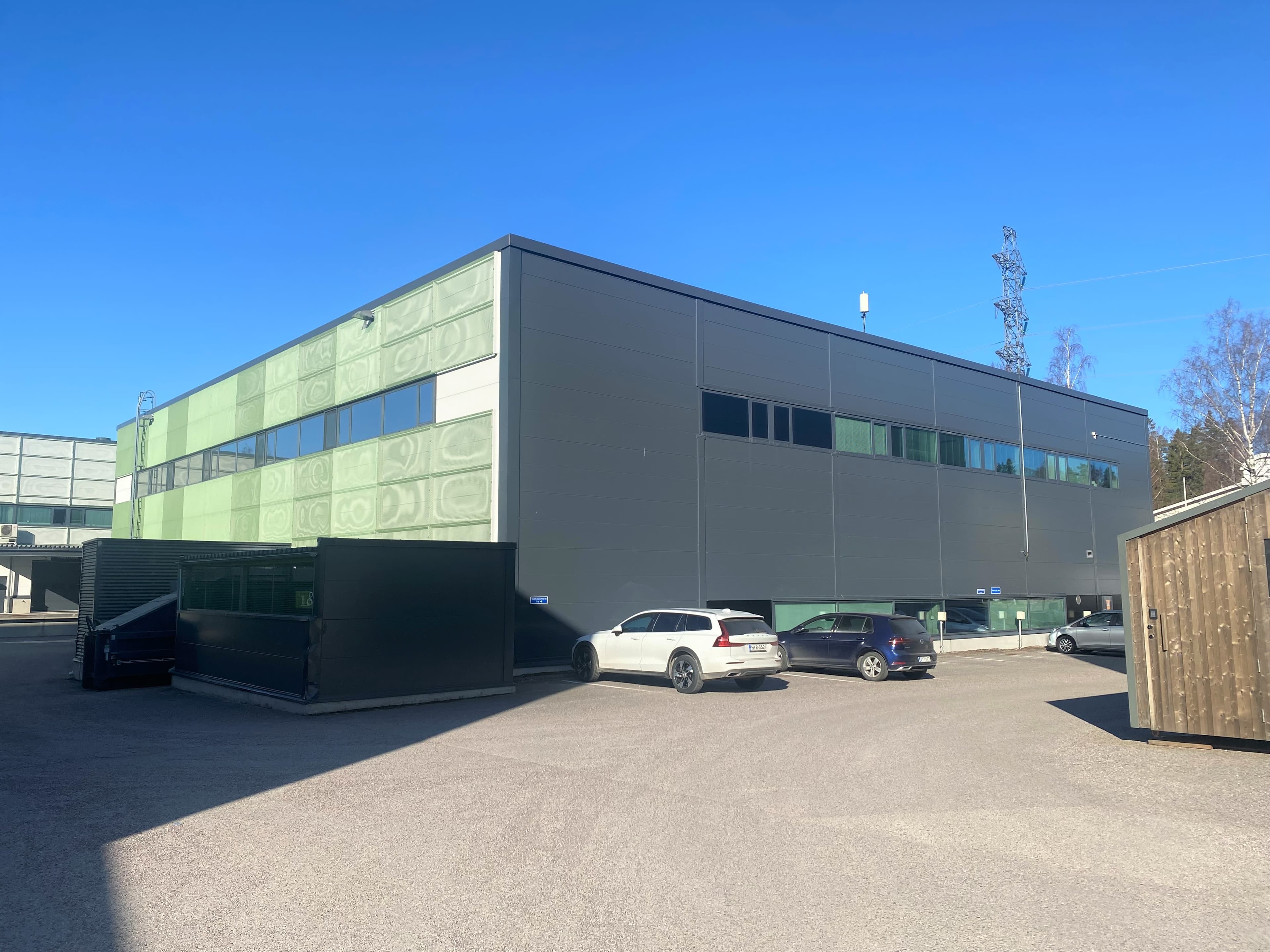 Moderni tummanharmaa ja vihreä rakennus sekä pysäköintialue aurinkoisena päivänä.