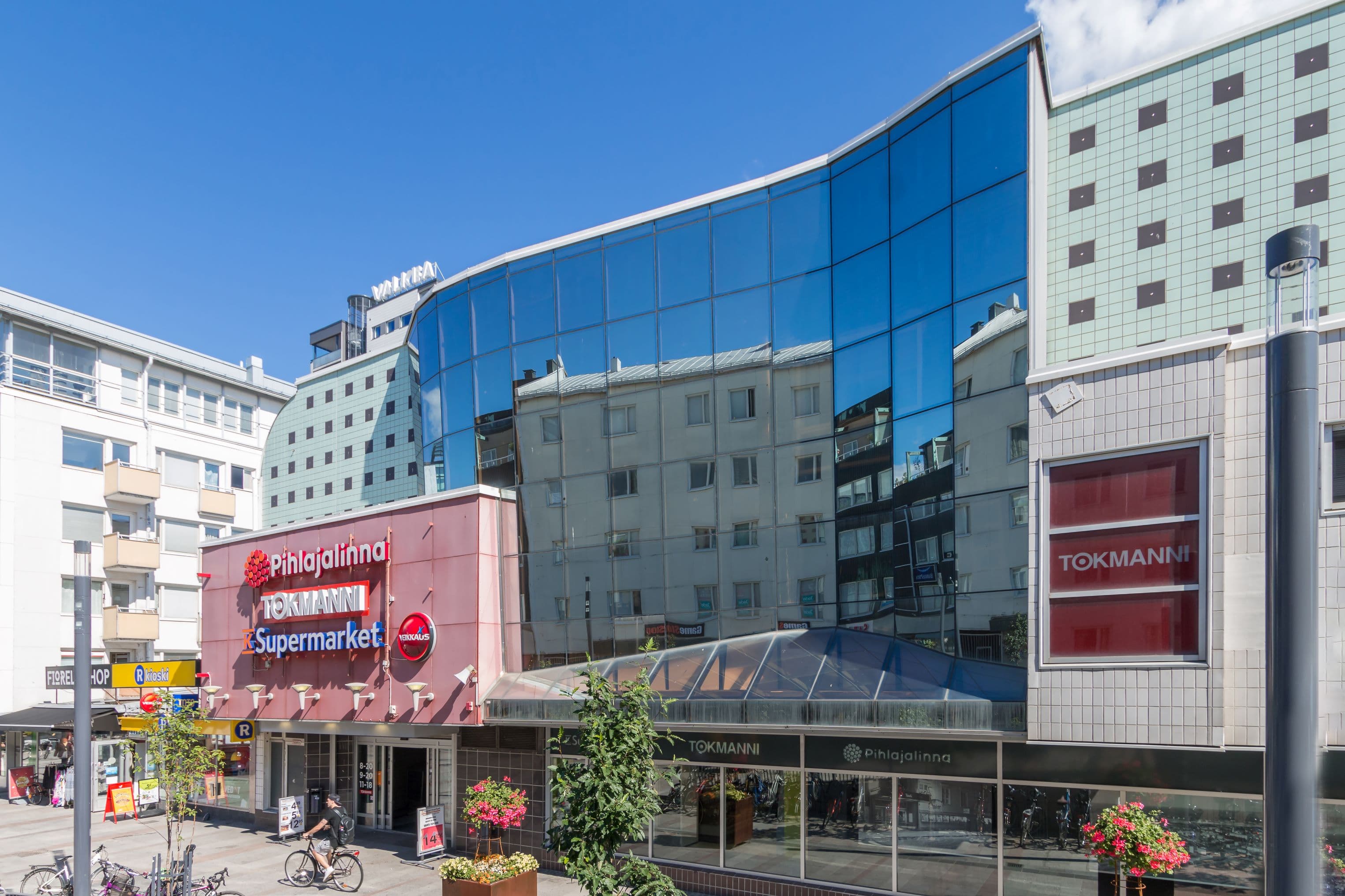Näkymä kadulta moderniin liikerakennukseen, jonka julkisivussa on Tokmannin, K-Supermarketin ja Pihlajalinnan kylttejä.