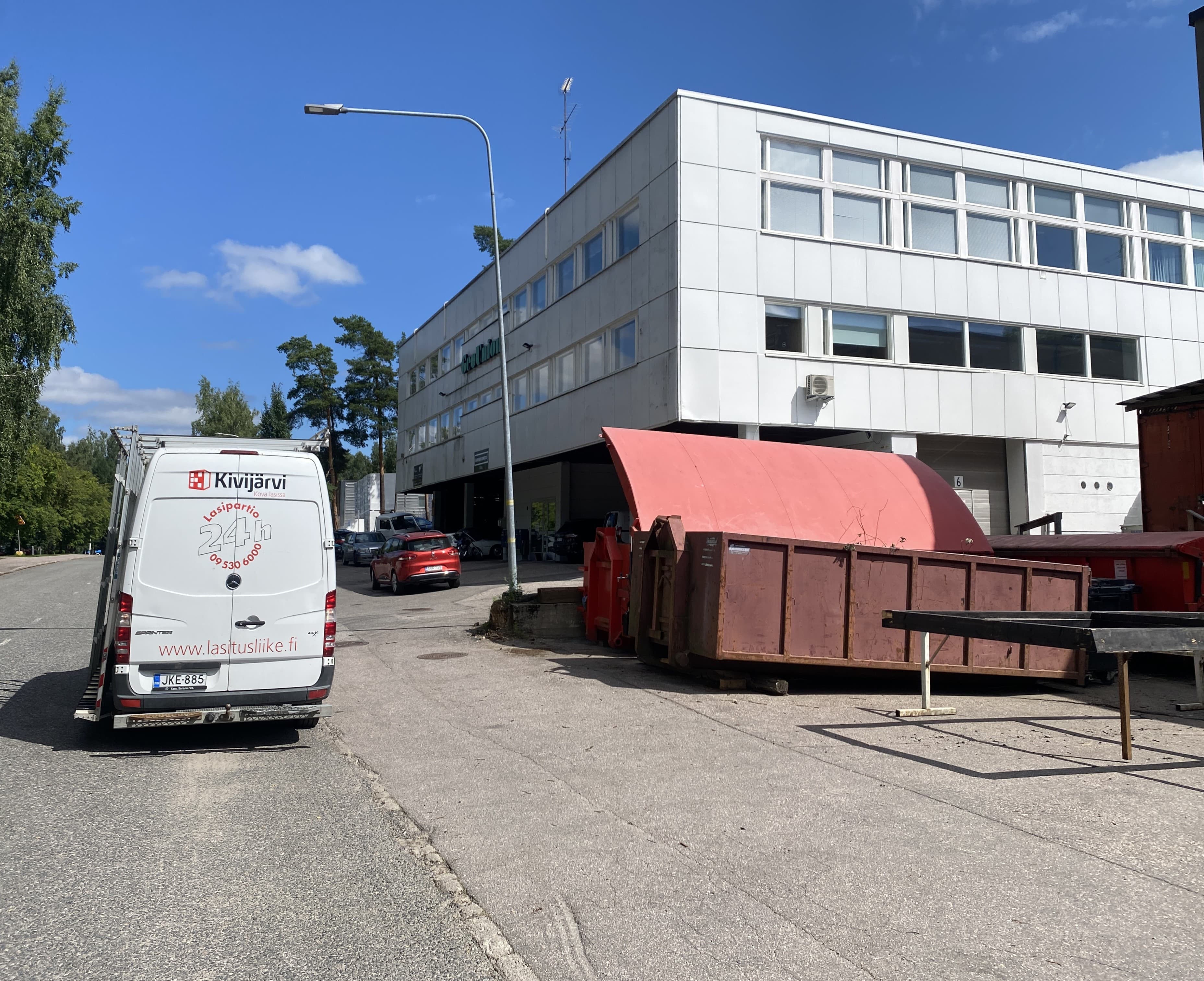 Valkoinen pakettiauto ja teollisuusrakennus, jonka edessä on suuri punainen siirtolava aurinkoisena päivänä.