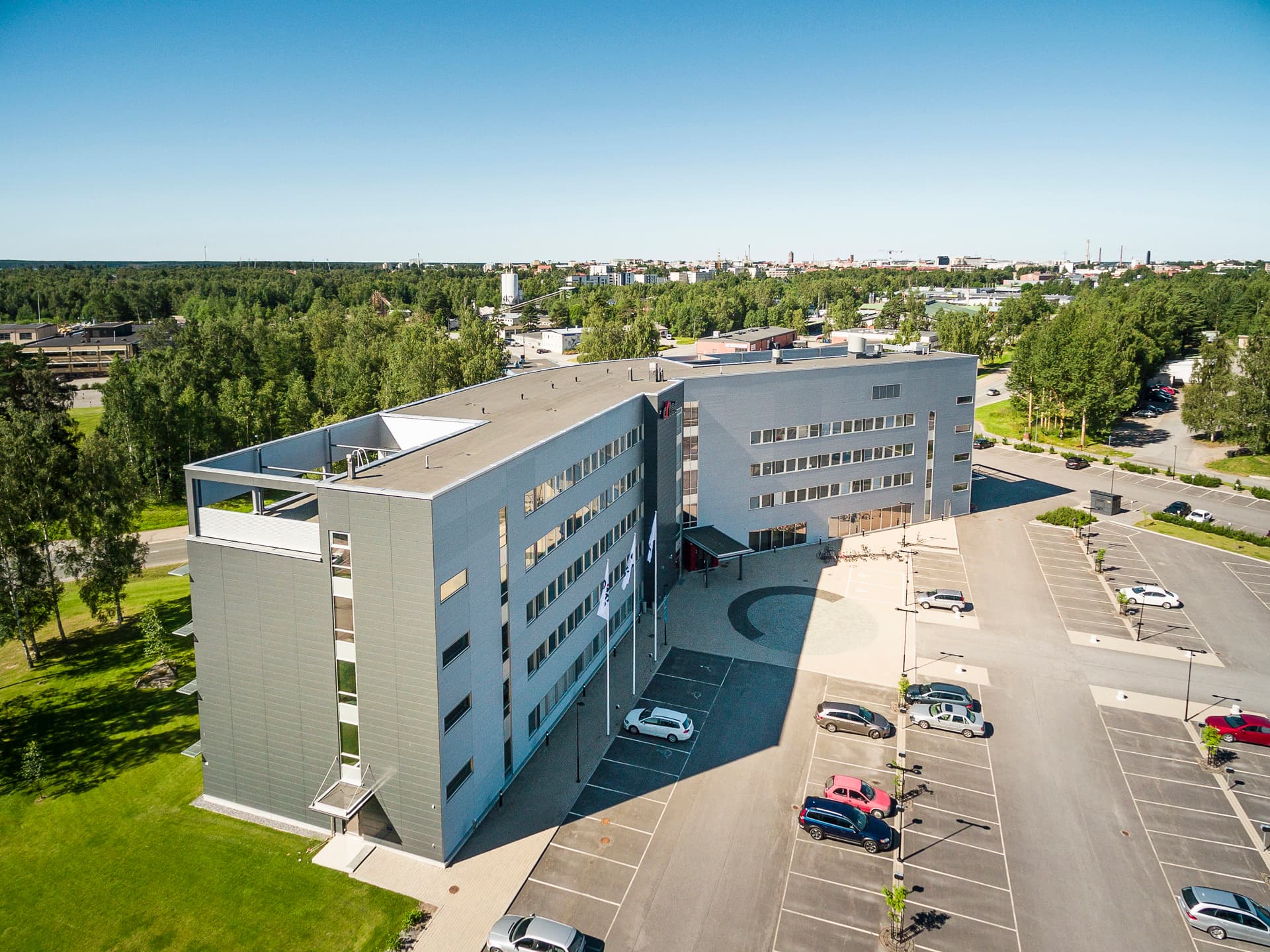 Ilmakuva modernista harmaasta toimistorakennuksesta ja sen pysäköintialueesta.