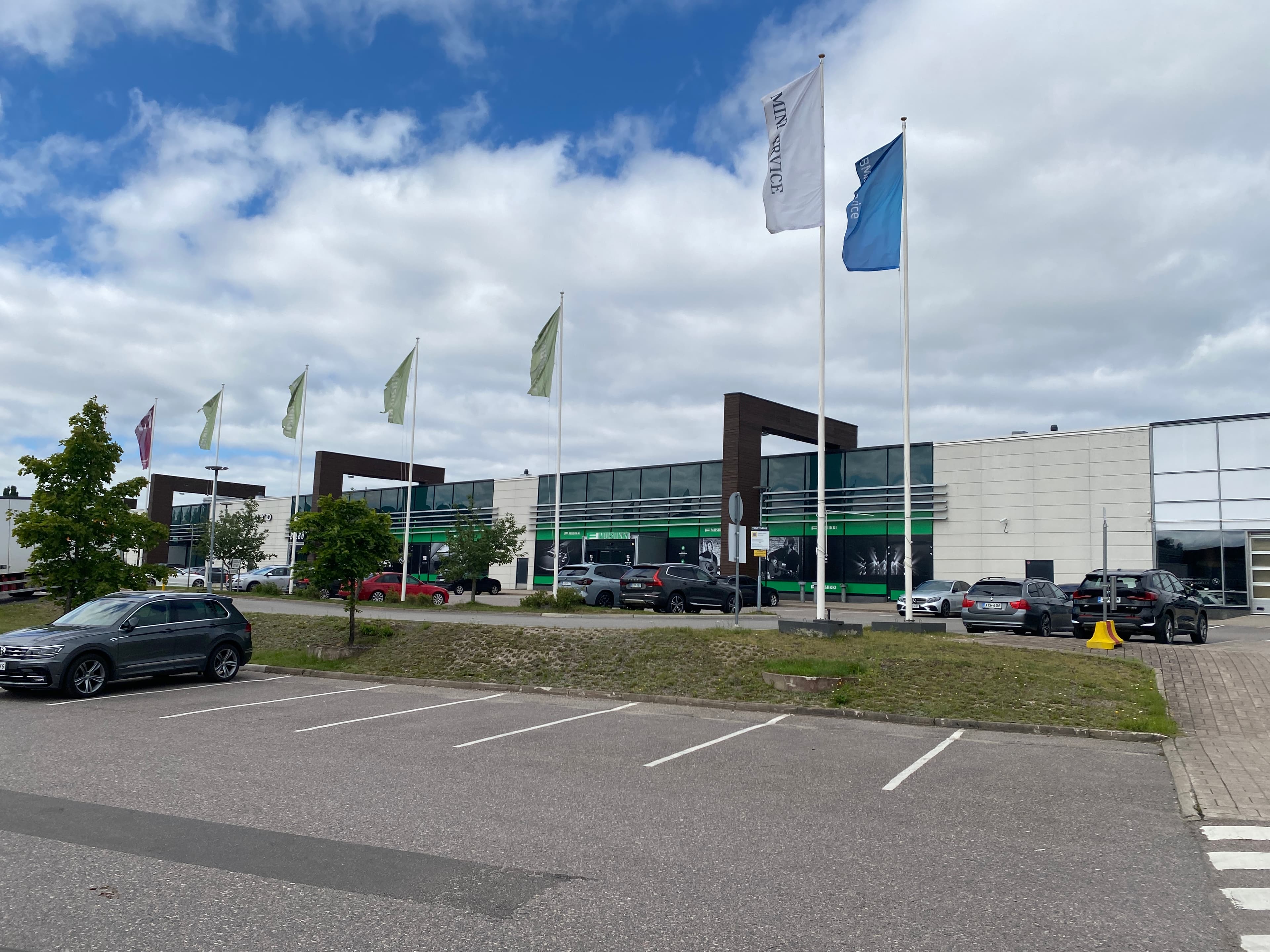 Näkymä liikerakennuksen edustalta, jossa on pysäköintialue ja lippusalkoja pilvisenä päivänä.