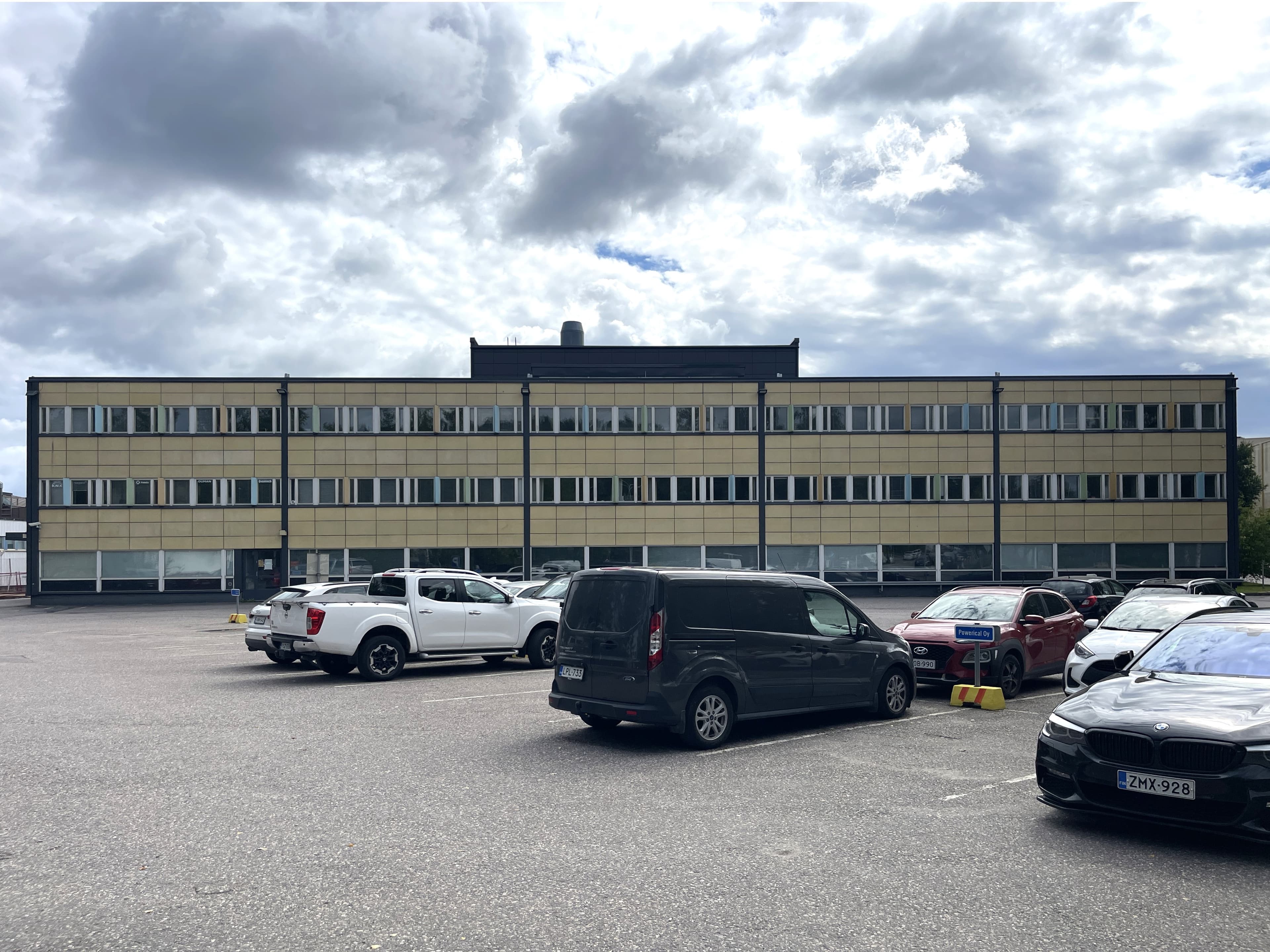 Kolmikerroksinen keltainen toimistorakennus ja sen edustalla oleva parkkipaikka autoineen pilvisenä päivänä.