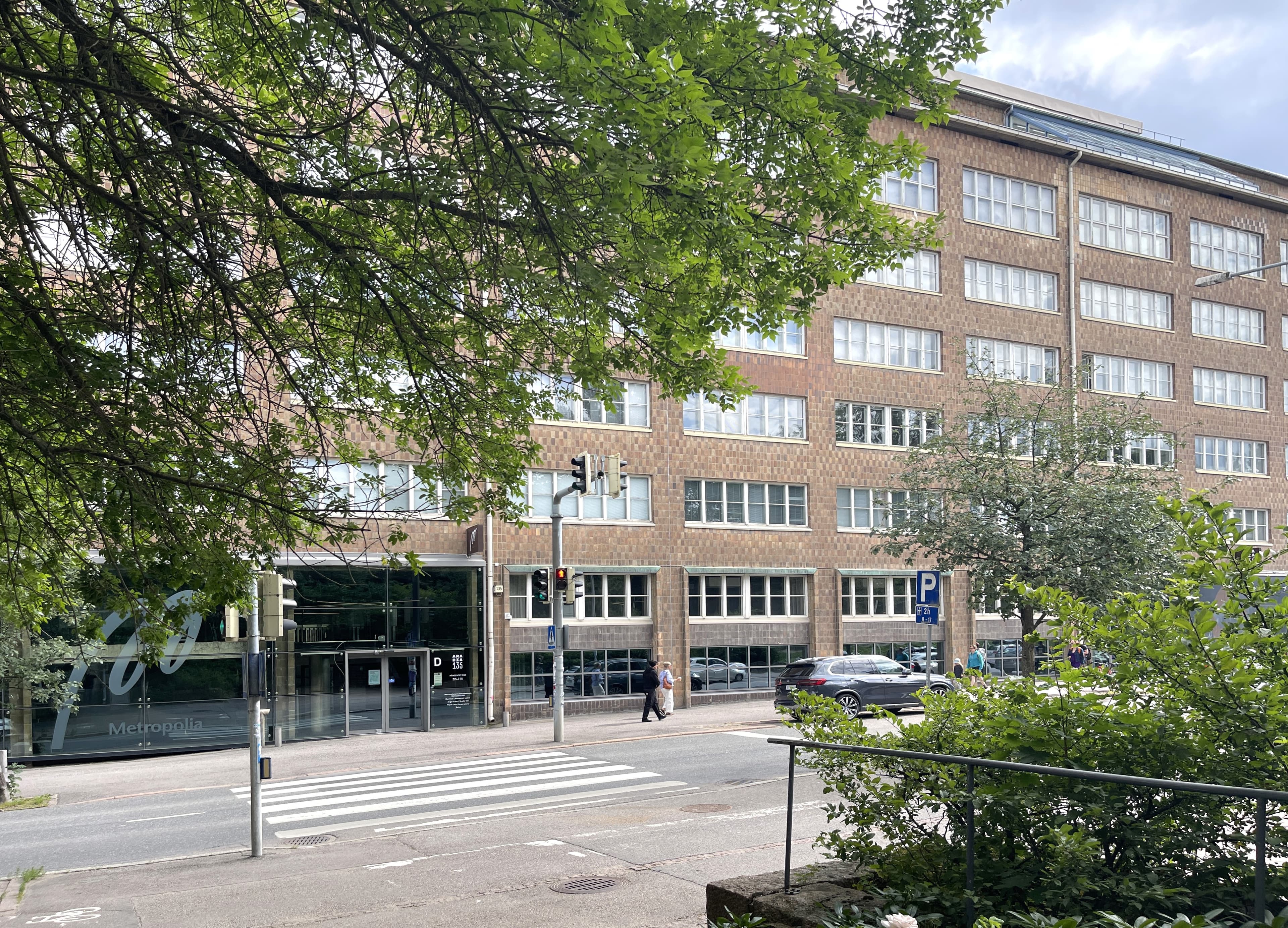 Näkymä suuresta tiilirakennuksesta ja kadusta puiden takaa.