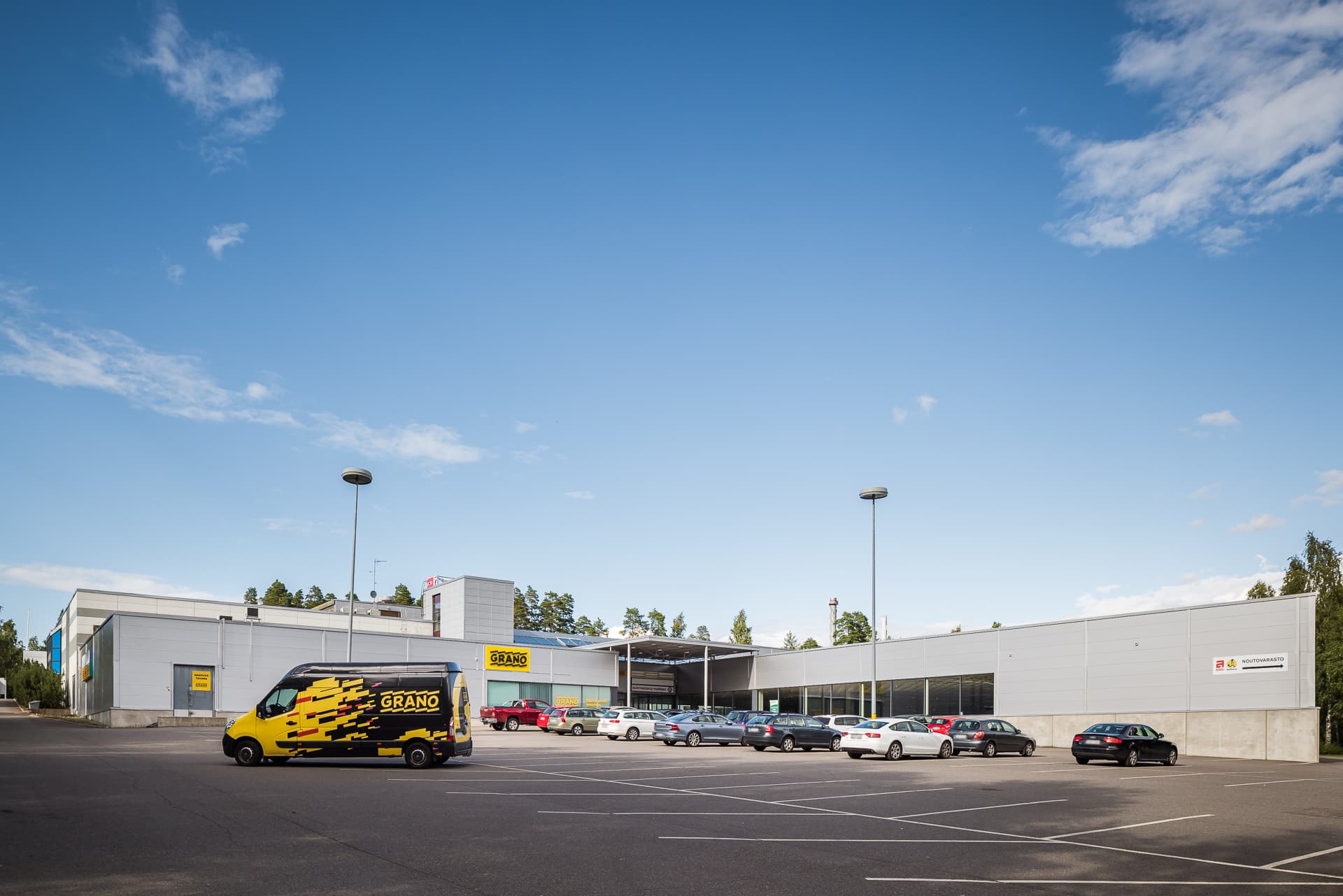 Moderni teollisuusrakennus ja sen parkkipaikka autoineen kirkkaana päivänä.