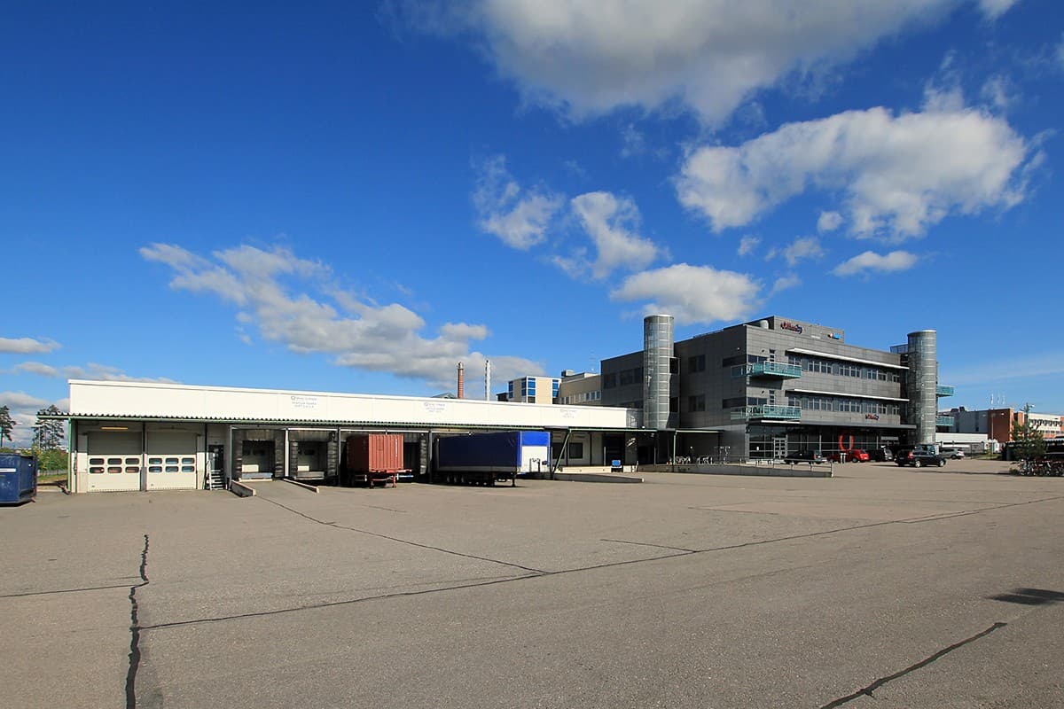 Teollisuus- ja toimistorakennus laajan asfalttipihan reunalla aurinkoisena päivänä.