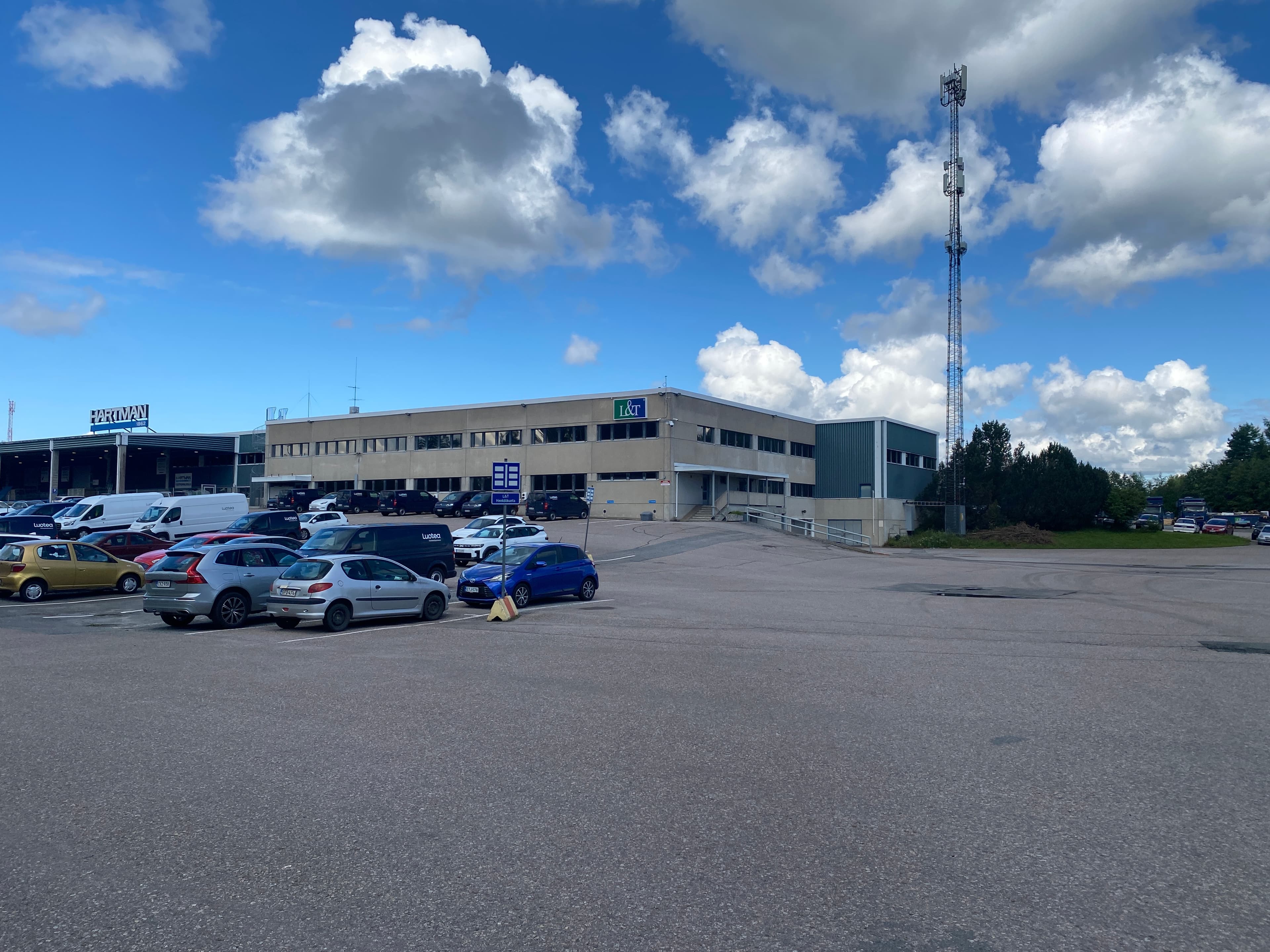 Näkymä liikerakennuksen parkkipaikalta, jossa on autoja ja korkea masto puolipilvisen taivaan alla.