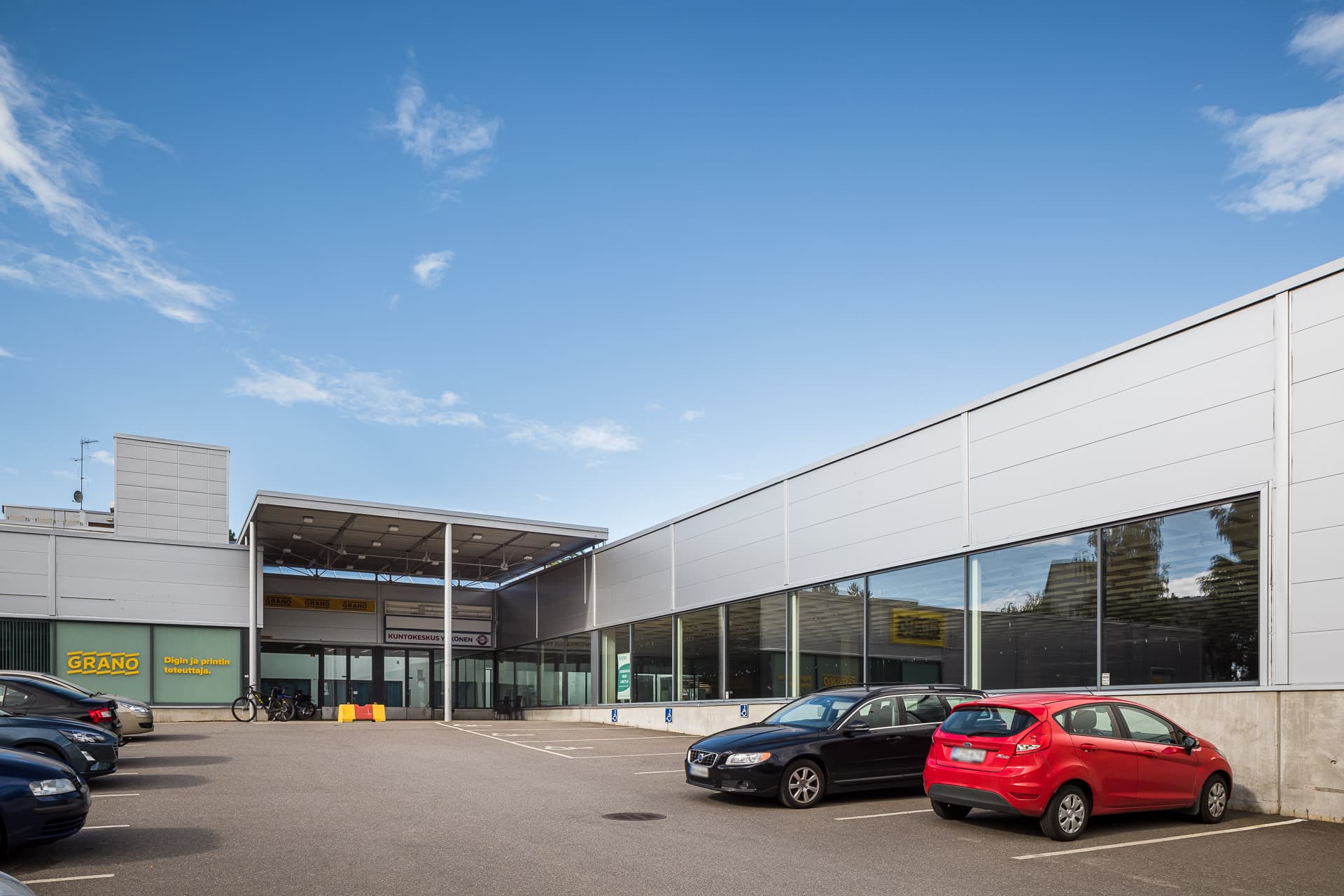 Moderni liikerakennus ja sen edessä oleva pysäköintialue autoineen aurinkoisena päivänä.