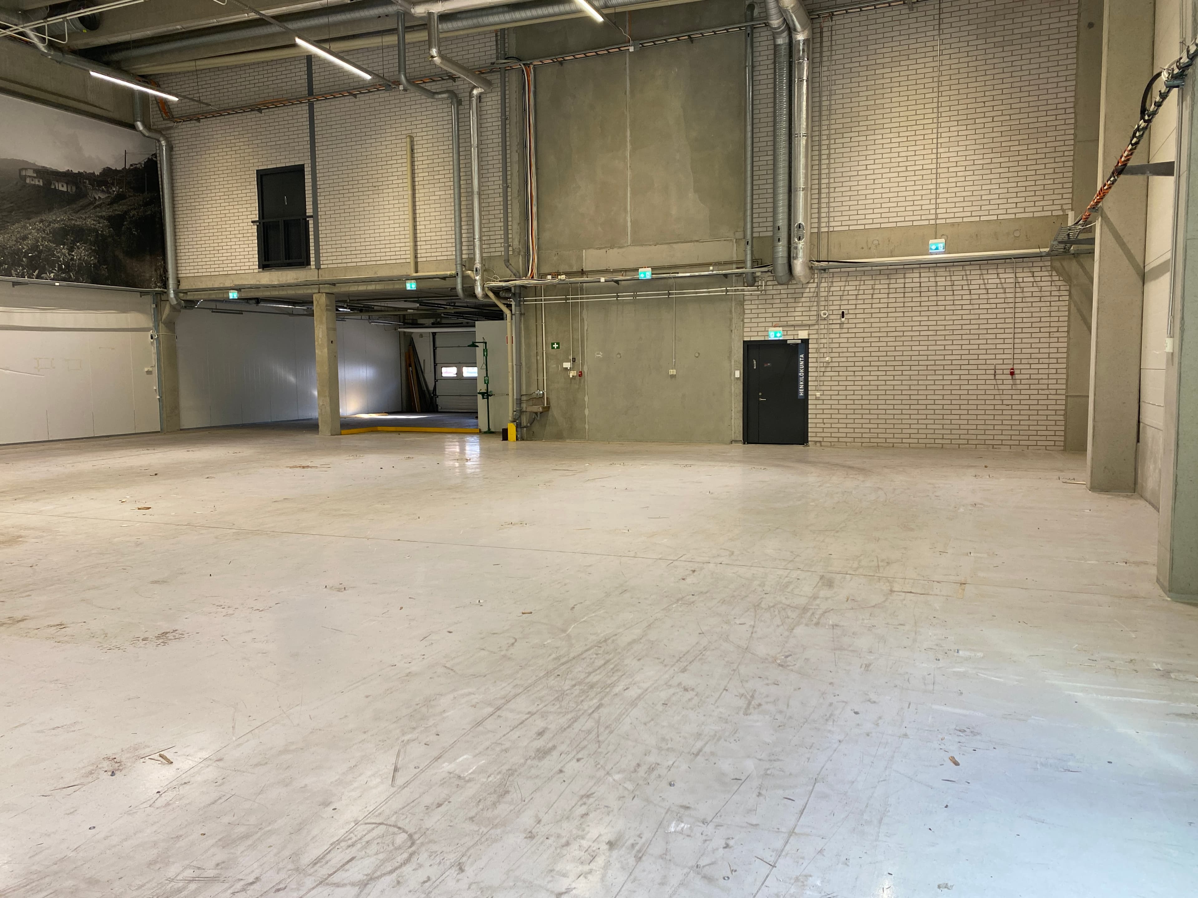Tyhjä ja avara teollisuushalli, jossa on betonilattia ja näkyvää putkistoa katossa.